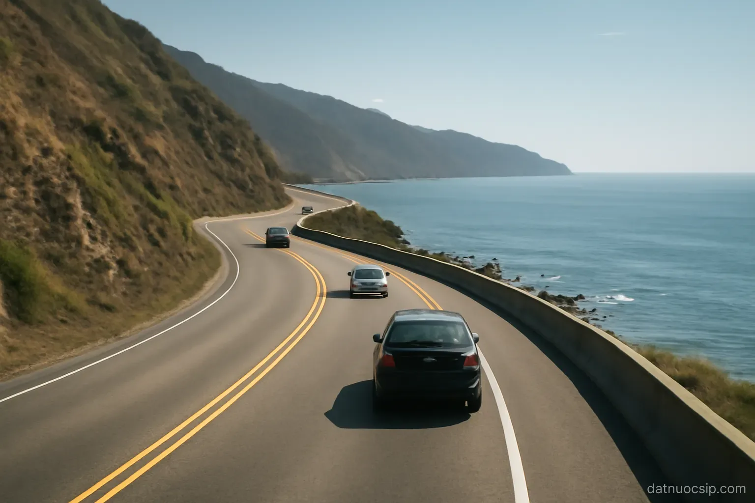 a coastal highway curving along a hillside with the sea below, few cars, clear day