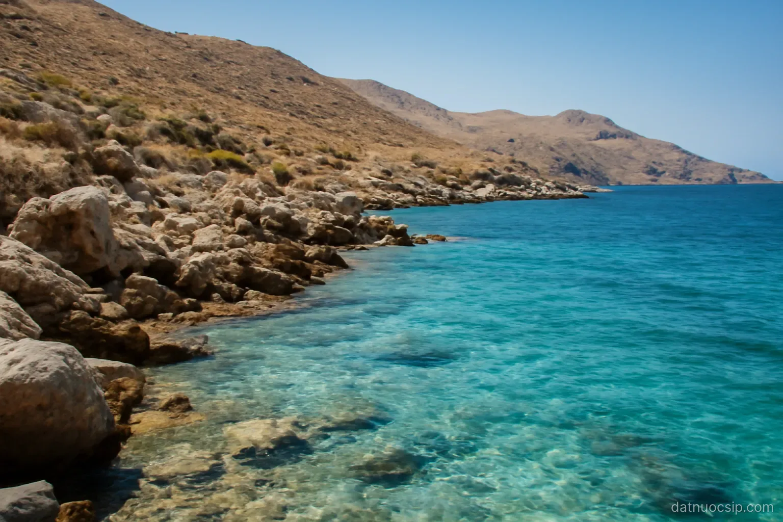 a rocky Mediterranean coastline with turquoise water and dry hills behind, wide view